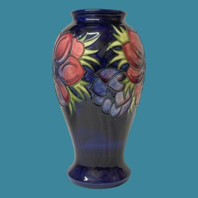 Photo of vase seen from an eye level with the sides of mauve anemones visible on the top left and right and a small blue unopened anemone at the centre.. The vase is set against a light air force blue colour which hi lights the deep blue base colour of the vase.