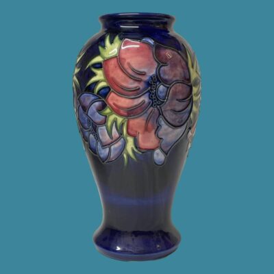 Main photo of vase seen from an eye level with a mauve anemone on display with part of the unopened small blue one to its left. Vase is set against a light air force blue colour which compliments & hi-lights the colours of the vase.