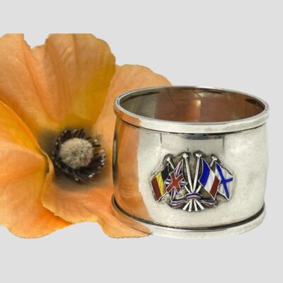 Main photo of napkin ring seen from a slightly raised eye level so part of the inside is visible. The ring is displayed next to a red poppy with the enamel flags i the foreground.