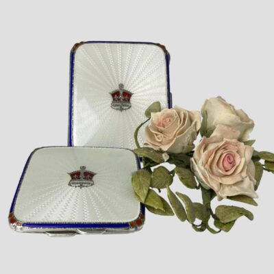 Main photo of both item displayed with the cigarette case upright on a stand and the compact laid flat below it and slightly to left. A small posy of pale pink roses is to the right of the compact. The lids f both items are mostly white guilloche enamel with a red and silver crown to the centre. The outer rim of the white enamel has royal blue enamel to the flat sides and red enamel to the corners giving a regal feel.