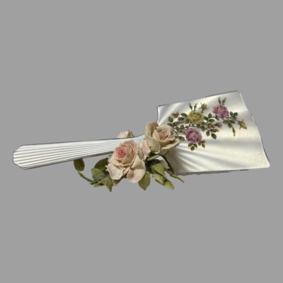 Main photo of mirror displayed with the mirror end on a stand and the handle out to the left. The enmel side of the mirror is on display with a small bunch of pale pink roses in the foreground as decoration.