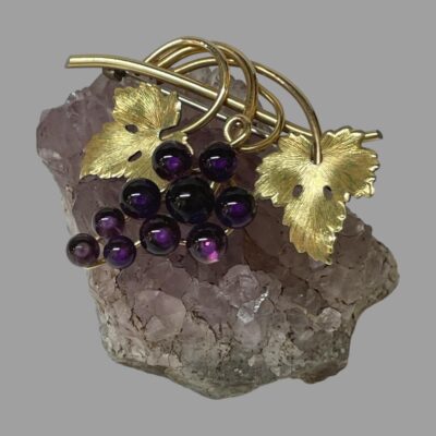 Main photo of the brooch on top of a small rock of natural amethyst. The photo is taken looking straight down at the brooch from above. The grapes are to the centre with a gilt vine leaf to either side. The amethyst stones making the grapes on the brooch are a dark purple in contract to the lilac of the rock. The gilt on the metal is in excellent condition and compliments the purple amethyst beautifully.