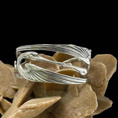 Main photo of cuff bracelet displayed on top of a large sand rose (formed desert sand) and shown with one of the cranes in full view at eye level and the beak of other visible. The bracelet is formed of 2 cut out crane figures placed completely mirrored one on left, one right and one upright one upside down and joined together. The centre of the bracelet is the heads with beaks joined together while the upper and lower bands are the cranes spread wings.