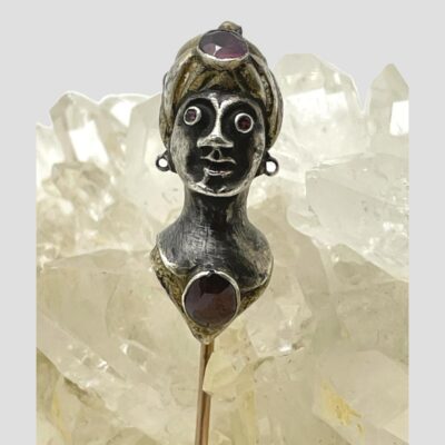 Main photo of pin leaning up against some quartz crystal with mostly just the head at the top of pin on show. The lady is facing straight at camera. The garnet cabochon necklace and amethyst to the top of her turban hat are visible as are the amethysts of her somewhat crazed looking eyes! The face and neck are silver and the turban hat and collar are in gilt.