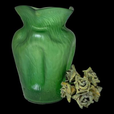 Main photo of vase seen from an eye level with one dimpled side in the foreground to the left. The vase is a striking slightly iridescent emerald green colour. There is a small posy of pale green coloured flowers on the righthand side of the vase.