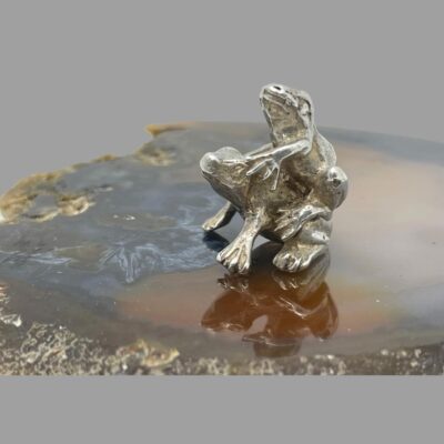 Main photo of the frogs displayed on top of a slice of agate. The frogs re both facing to the left with one piggy-backing on the other. Both frogs are facing the same way, in this shot left.
