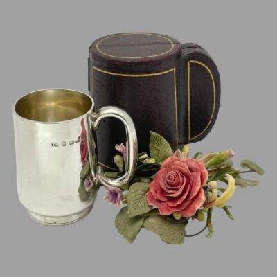 Main photo of christening displayed outside and to the left of its case which is closed. The handles for both the mug and case are on the right and there is a small posy of flowers including a red rose to the right of the mug and in front of the case.
