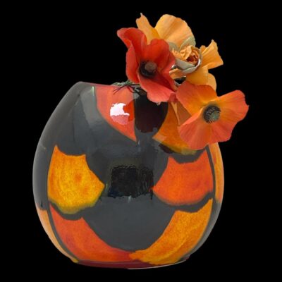 Main photo of vase seen from the patchwork colour side. The background colour is black with blue and a handful of patches in light and dark orange spaced out mostly around the outer edge. The patches are not uniform in size. There is a small posy of poppies in red and orange inside and to the right of the vase - their colours are almost a perfect match for the orange patches on the vase. The vase is set against a black background.