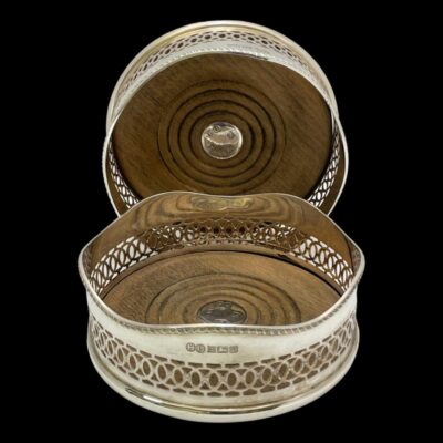 Main photo of the 2 coasters shown from a slightly raised angle looking diagonally down. One is in the background on its side to show the inside wood area and the other is in the foreground laid flat. The hallmark is visible on the outer rim of one in foreground and the hallmark on the one in background is visible to the disc at the centre.