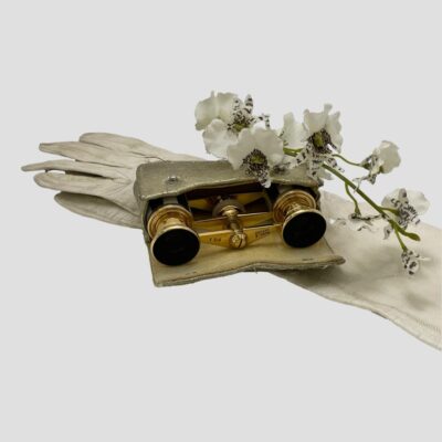 Main photo of opera glasses displayed inside their case. The case is laid flat with the cover open displaying part of the glasses which are slightly protruding out. The case is on top of a pair of long off white gloves with a small sprig of orchid flowers draped over the top.