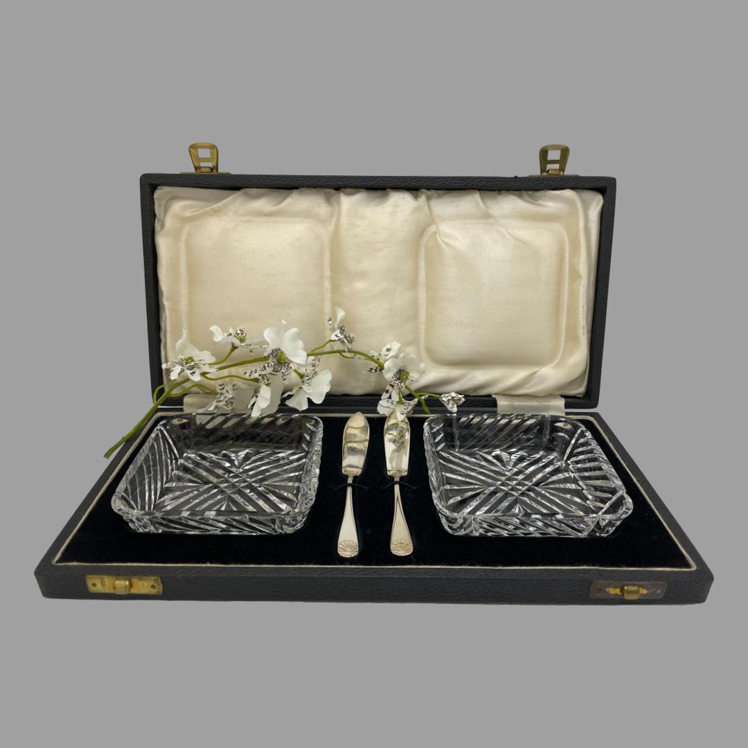 Main photo showing the case open with all pieces displayed in their fitted compartments. The butter dishes sit in a shallow dip on the left and right while the knives sit under elastic to the centre. The lid is upright with the silk interior showing and there is a sprig of white orchids across the centre.