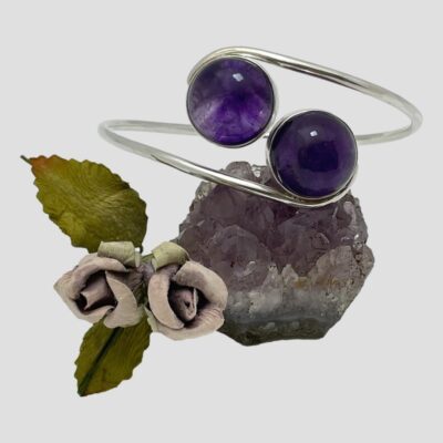 Main photo of bracelet displayed with the amethysts in the foreground and balanced on top of a small rock of natural amethyst. The amethysts on the bracelet are a deeper shade of purple than the natural rock. There is a sprig of pale smoky purple paper roses to the left.