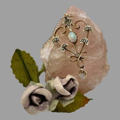 Main photo of necklace displayed over a rock of rose quartz. The pendant is on full display with oval shaped opal to the centre of the almost heart shaped frame with a smaller round opal dangling at the bottom. Two small wing shaped gold sections are to either side of the oval opal with seed pearls as well as an almost star shaped section which is attached to the bail. The chain is only just visible. There are 2 small light smoky purple coloured faux roses to the bottom left of the quartz.