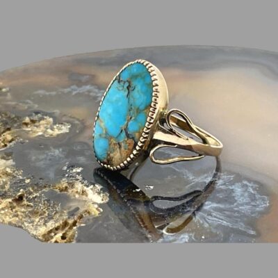 Photo of ring displayed on a slice of natural agate with the ring front facing left displaying the unusual shoulder of 2 elongated loops of gold one above and one below the main ring band. The looped end is near the stone setting .