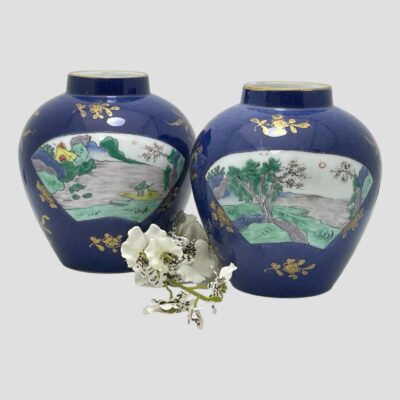 Main photo of jars shown side by side with one slightly to the foreground than other. The one in front shows just the river scene with pine trees and other slightly to the back has river scene with a fisherman in a yellow boat to the right and a yellow and red tent to the left. Each vase has the same scenes within a fan shaped window. There is a spray of small white orchids in between the 2 jars.