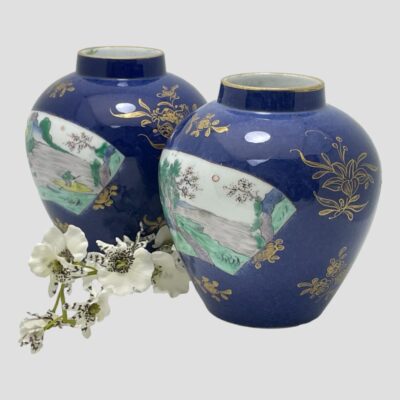 Secondary photo of the 2 jars shown side by side from a slightly raised and side on angle so one vase appears in the background top left and one in foreground is in the bottom right. There is a spray of small white orchid flowers in between the 2 jars.