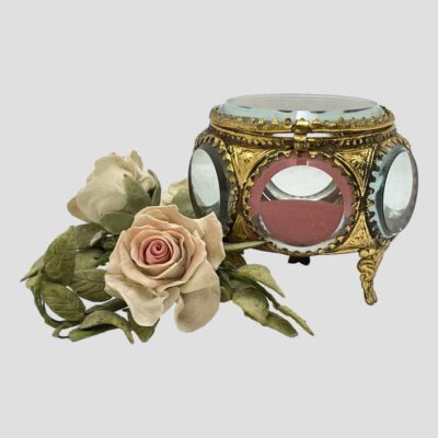 Main photo of box seen from an eye level and seen displayed with a small posy of pale pink roses to its left. The box is right of the roses with one round window to the centre and windows to left and right partially visible. The red (or dusky dark pink) of the padding is also visible inside the casket.