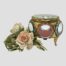 Main photo of box seen from an eye level and seen displayed with a small posy of pale pink roses to its left. The box is right of the roses with one round window to the centre and windows to left and right partially visible. The red (or dusky dark pink) of the padding is also visible inside the casket.