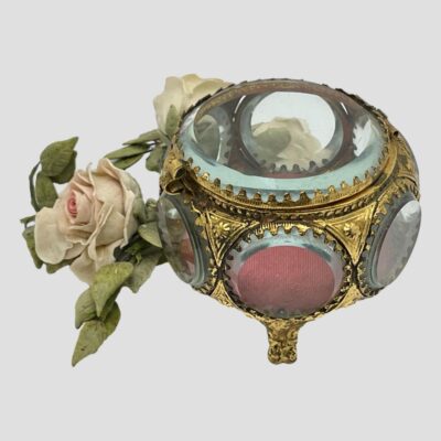 Secondary photo of box seen from a slightly raised angle looking diagonally down. The clear bevel edged glass is visible to the top with window to the front showing the red (dusky dark pink) cushion. The mirror to the back is also shown in full but through the lid glass. The small posy of pale pink roses is displayed to the back of the box.