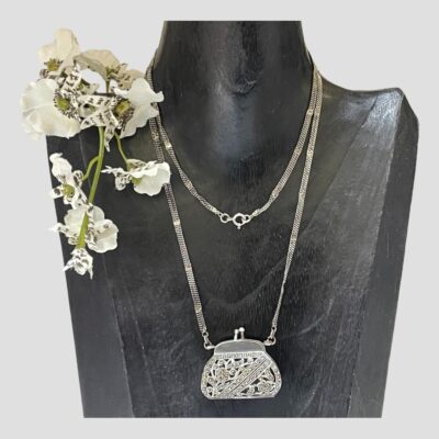 Main photo of necklace with attached purse pendant displayed on a dark wooden display stand. The chain is long and has had to be double round the neck of the display. The purse pendant is a nice size and is a very visible statement. The is a spray of small white orchids on the left shoulder of the display stand.