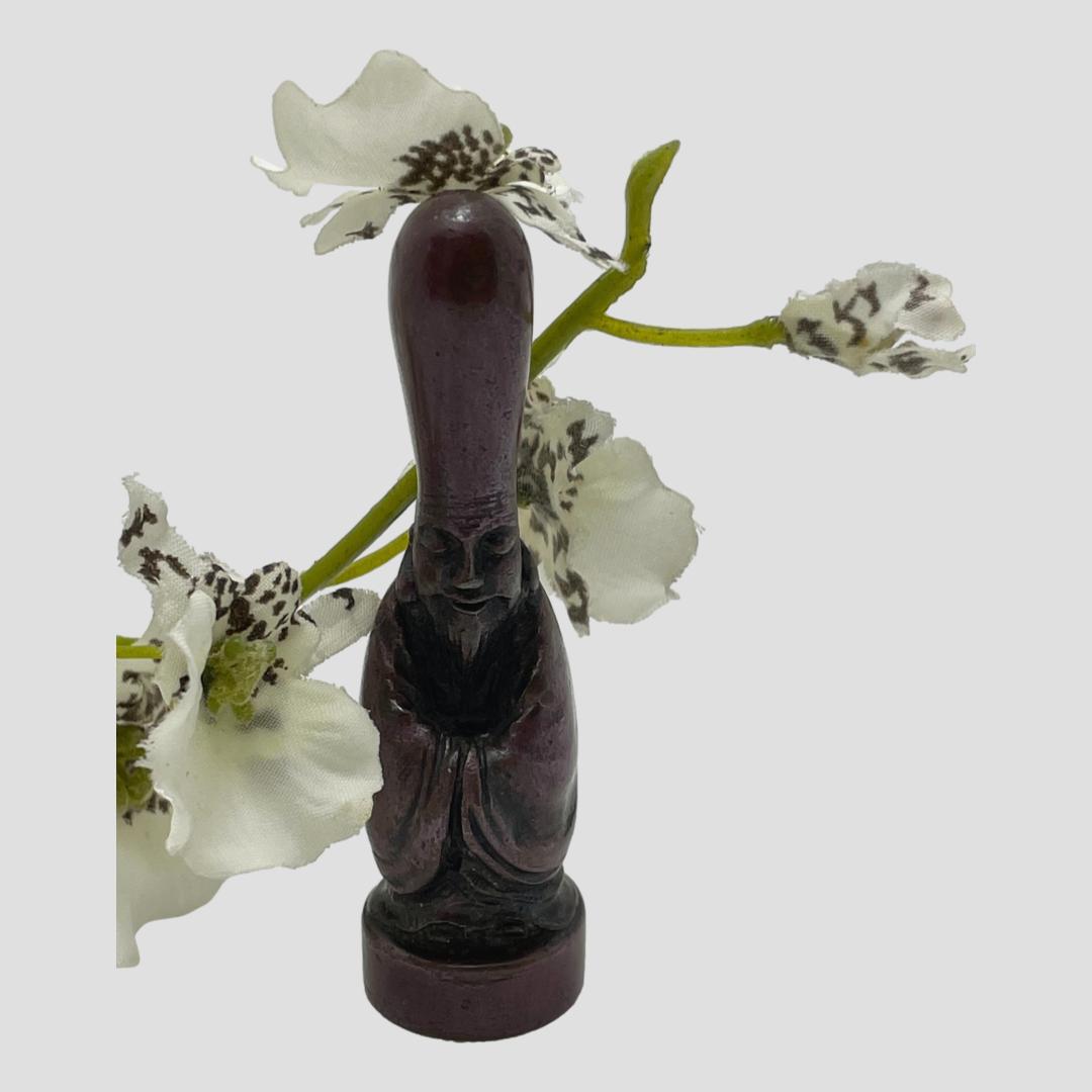 Main photo showing Fukurokuju deity forward facing and from a very slight height. He has both hands inside opposing arms of his haori kimono and has a lovely serene and calming face. His elongated head is almost the size of the rest of his body. There is a spray of small white orchids behind the okimono giving an extra Oriental feel!