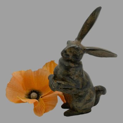 Close up shot of the hare seen from a slightly raised angle. The rabbit is now posed with the front paws facing to the bottom left corner and the upright ear pointing to the top right corner. The poppy has been moved to behind the hare near the front end.