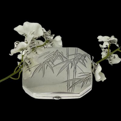 Main photo of compact laid on a flat surface against a black background. The photo is taken from a little height looking down diagonally at the bamboo decoration on the lid. The compact is propped up slightly at the back with a spray of small white orchid flowers.