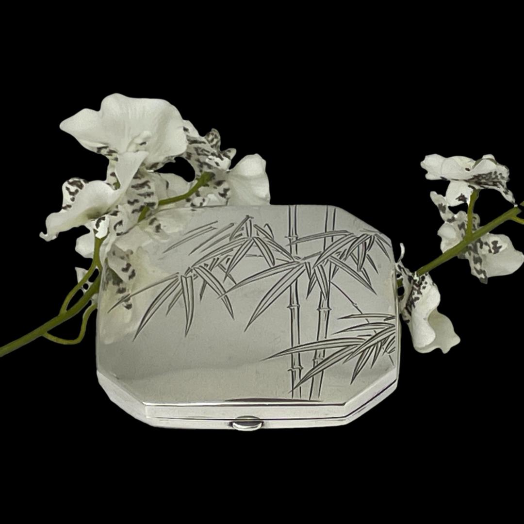 Main photo of compact laid on a flat surface against a black background. The photo is taken from a little height looking down diagonally at the bamboo decoration on the lid. The compact is propped up slightly at the back with a spray of small white orchid flowers.