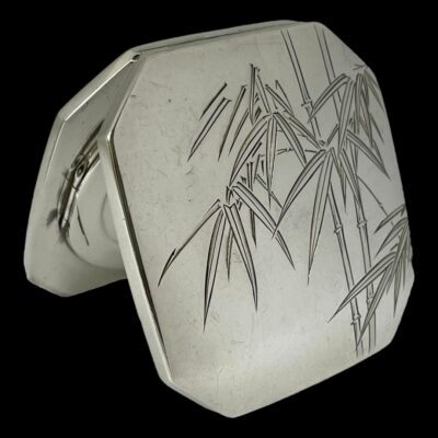 Photo of compact shown open and propping itself up with the lid. The top side of compact with the larger decoration of etched bamboo is shown.