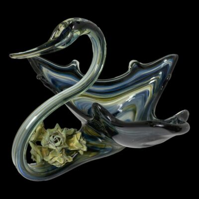 Main photo of swan shaped bowl with the front of the swan in the bottom left and the bowl back end tapering to a point in the top right corner of photo. The bowl is a triangular shape giving an impression of a swan with wings outstretched. The overall colouring is multiple shades of blue stripes in clear glass with a golden accent lower in the bowl. The is a small posy of pale green/blue roses on the base of the neck.