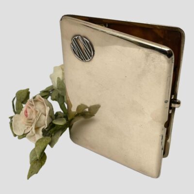 Photo of cigarette case shown upright and slightly ajar. The MGM logo is to the top left and a pale pink rose is to the bottom left. The angle of shot is slightly raised so we are looking downward from the top.