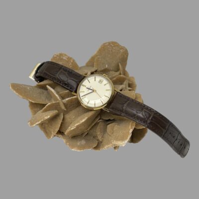 Main photo of the watch displayed laying across a rock of desert sand rose with the buckle end on the left and end of strap on the right. The watch face is to the centre of shot .