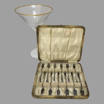 Main photo showing the forks inside the fitted case with the lid open upright and leaning against a cocktail glass.