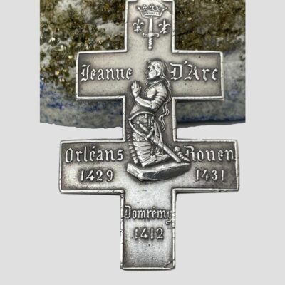 Close up photo of the front of cross with an image of Joan of Arc knelt in prayer to the centre, coat of arms to the top and the 3 significant dates of her short life ; birth year and place, Orleans 1429 and 1431 the year of her execution at the stake.