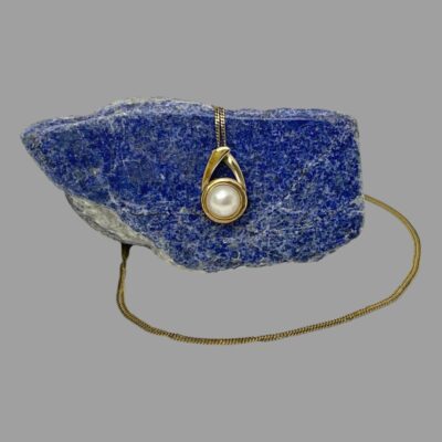 Main photo of necklace with pendant displayed hanging off the side of a rock of natural lapis lazuli, the blue of the lapis enhances the pearl and gold. The chain goes down the back of the lapis rock and reappears out front in a circular pattern with the end disappearing behind the rock.