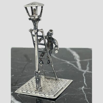 Main photo showing miniature with the lamp in the bottom left corner of shot. The piece is shown diagonally displayed with lamp in left foreground, ladder leaning against it with the lamplighter halfway up the ladder. The piece is displayed on top of black marble with white line inclusions.