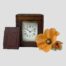 Main photo of clock with the leather piece removed from the outer case displaying the clock face thru a window. The leather window cover is leaning against the case on the left and a red poppy is laid to the right of the clock.