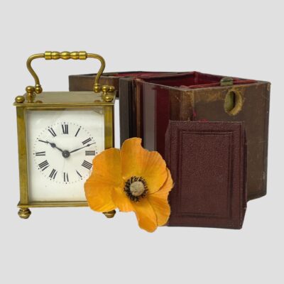 Photo of clock displayed outside of its case and set to the left. The empty leather case, on the right has the window piece removed and placed in front. A single red poppy is placed in the centre between the clock and the case.