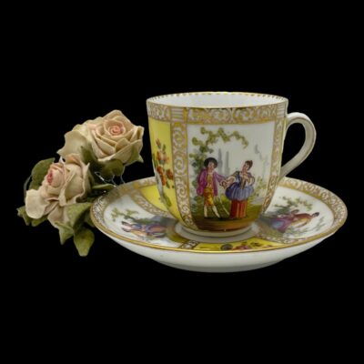 Main photo of cup on top of saucer seen from a near eye level. The handle is to the right. One Hand-painted panel with courting couple is on display to the centre of the cup (walking hand in hand) and 2 panels with courting couples are visible to left and right on the saucer. There is a small posy of pale pink roses to the left.