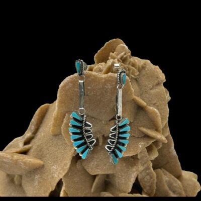Main photo of earrings displayed hanging from a rock of desert sand rose. The earrings are forward facing. There is a single turquoise stone to the top on the front of the posts with a flat piece of silver hanging down from here and a fan like shape piece to the ends each with 7 turquoise stones .