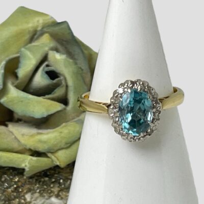 Main photo of ring displayed on a cone shaped white ring display stand with the ring front forward facing. There is a small gold flecked rock to the left of the cone with a small green/blue rose to its left.