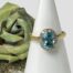 Main photo of ring displayed on a cone shaped white ring display stand with the ring front forward facing. There is a small gold flecked rock to the left of the cone with a small green/blue rose to its left.