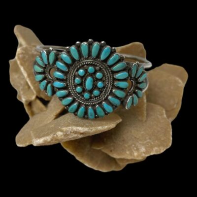 Main photo of bracelet displayed on top of a rock of desert sand rose with the front facing forward. There is a large circle to the centre with a frame of needlepoint turquoise all facing inward, a gap of silver with rope like pattern, then another circle of round turquoise stones with an oval to the centre. The centre circle is flanked to either side by a semi circle of 7 needlepoint turquoise stones. The open end of the semi circles are attached to the central circle.