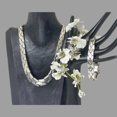 Main photo showing the necklace displayed on a dark wood bust and the bracelet hanging from the fingers of a dark hand display stand. The necklace is shown forward facing and then bracelet is seen width ways. There is a spray of small white orchids between the 2 stands.