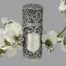Main photo of scent bottle seen with the empty cartouche forward facing. Apart from the cartouche which is plain flat silver, the rest of the bottle is intricately embossed with swirls and flowers. There is a spray of small white orchids in the background.