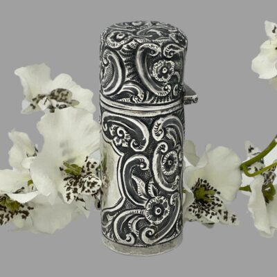 Photo of scent bottle seen from a side angle with the cartouche front facing left. There is still a spray of small white orchids in the background.