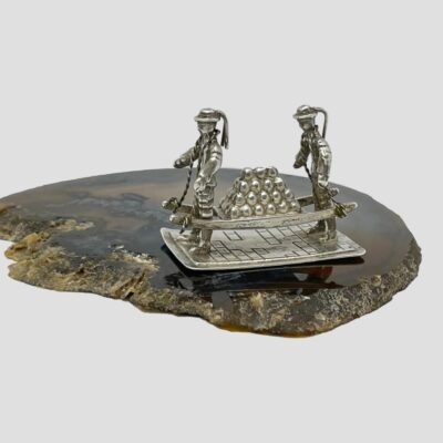 Main photo of miniature seen from an almost eye level sidelong angle with the sailors facing towards left. The cannon balls are stacked into a pyramid shape on top of the hand held barrow. The sailors are both hands free and the barrow is raised on the rope around the necks only. The miniature is on top of a slice of natural agate.