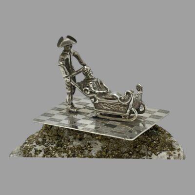 Main photo of miniature with the sleigh facing right. The man pushing the sleigh is wearing a tricorn hat. The sleigh is quite a fancy affair and the passenger looks very comfortable inside.