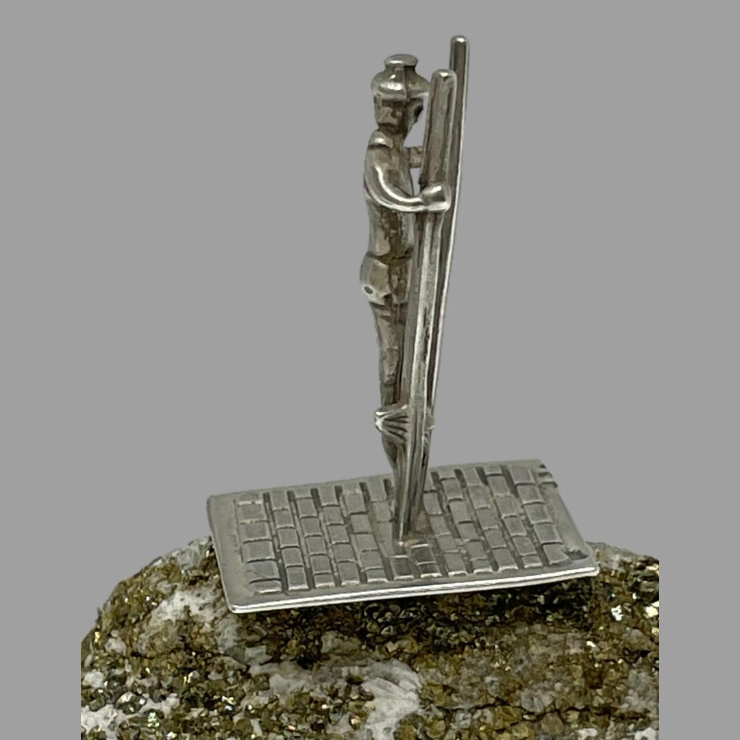 Main photo of stilt walker shown in profile facing direct to the right. The miniature is on top of a small natural rock. The stilts are not high ones with the standing ledge low to the ground so the walker is holding the top of the stilts.