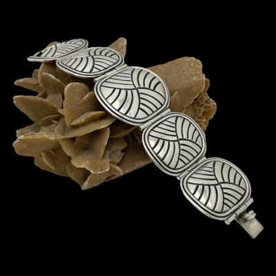 Main photo of bracelet displayed lying across a rock of desert sand rose diagonally. The clasp is undone so the full length of the bracelet is on display with the largest centre panel tilted at the top of the rock. The clasp fastener end is in the bottom right corner . The sand rose and bracelet are set against a black background which enhances the bracelet.